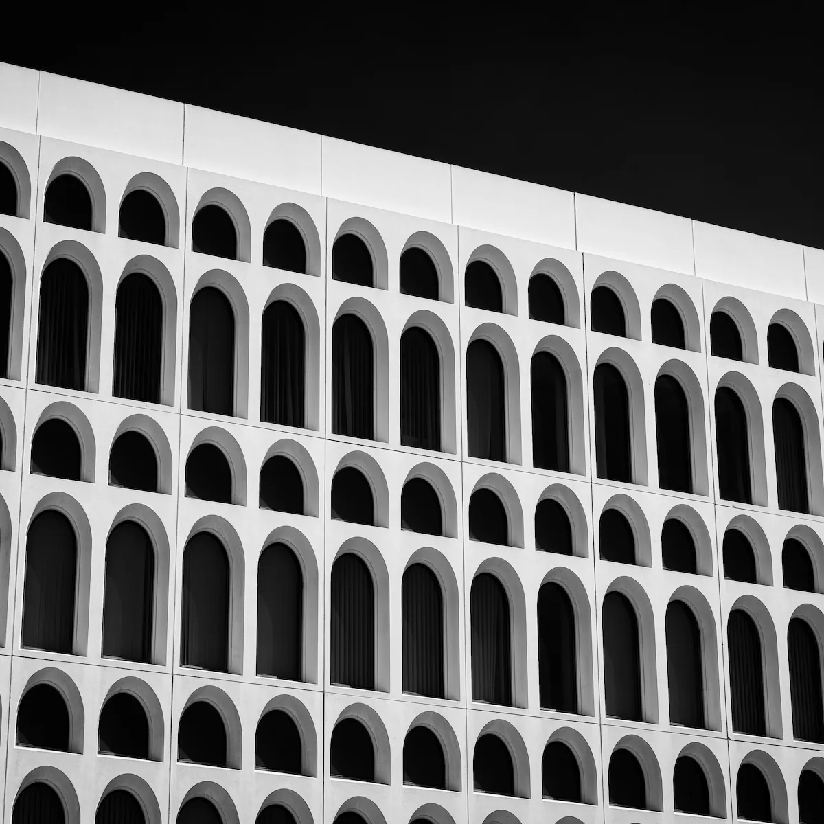 A white precast concrete facade seen from below at a sharp angle, its arched window openings stacked in rows that recede across the frame. Hard sun leaves the concrete near-white and the recessed windows black, against a sky rendered almost as dark. Black and white.