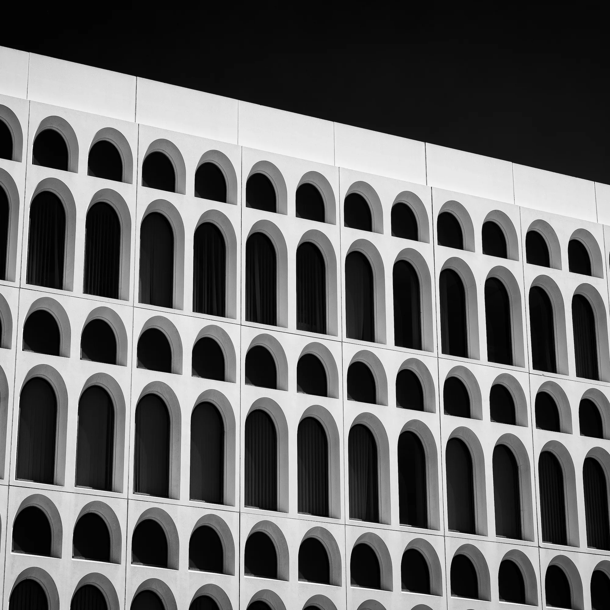 A white precast concrete facade seen from below at a sharp angle, its arched window openings stacked in rows that recede across the frame. Hard sun leaves the concrete near-white and the recessed windows black, against a sky rendered almost as dark. Black and white.