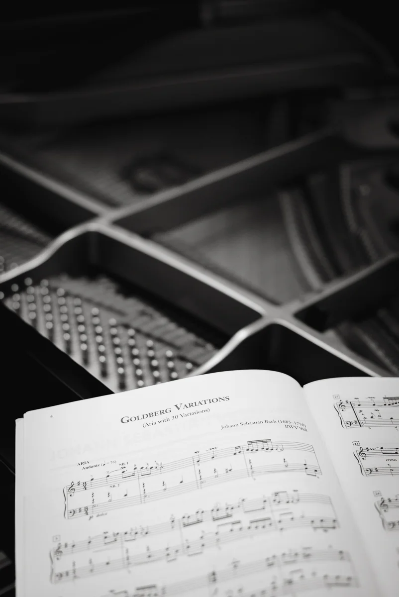 An open score of Bach's Goldberg Variations resting inside a grand piano, the strings, dampers and cast-iron frame out of focus behind it. Black and white.