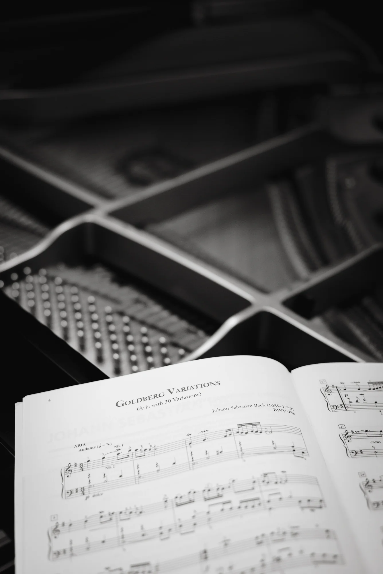 An open score of Bach's Goldberg Variations resting inside a grand piano, the strings, dampers and cast-iron frame out of focus behind it. Black and white.