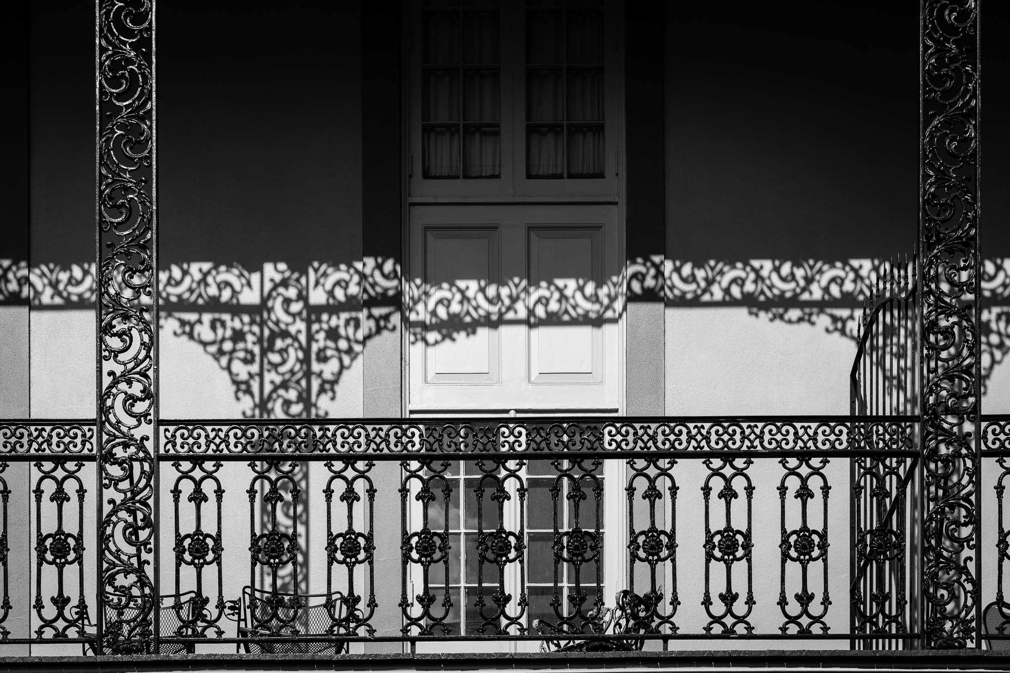 A cast-iron balcony seen straight on, its railing and scrolled panels worked in dense floral patterns. Low sun prints the ironwork's shadow across the pale wall behind it, doubling the pattern beside a tall shuttered door. Black and white.