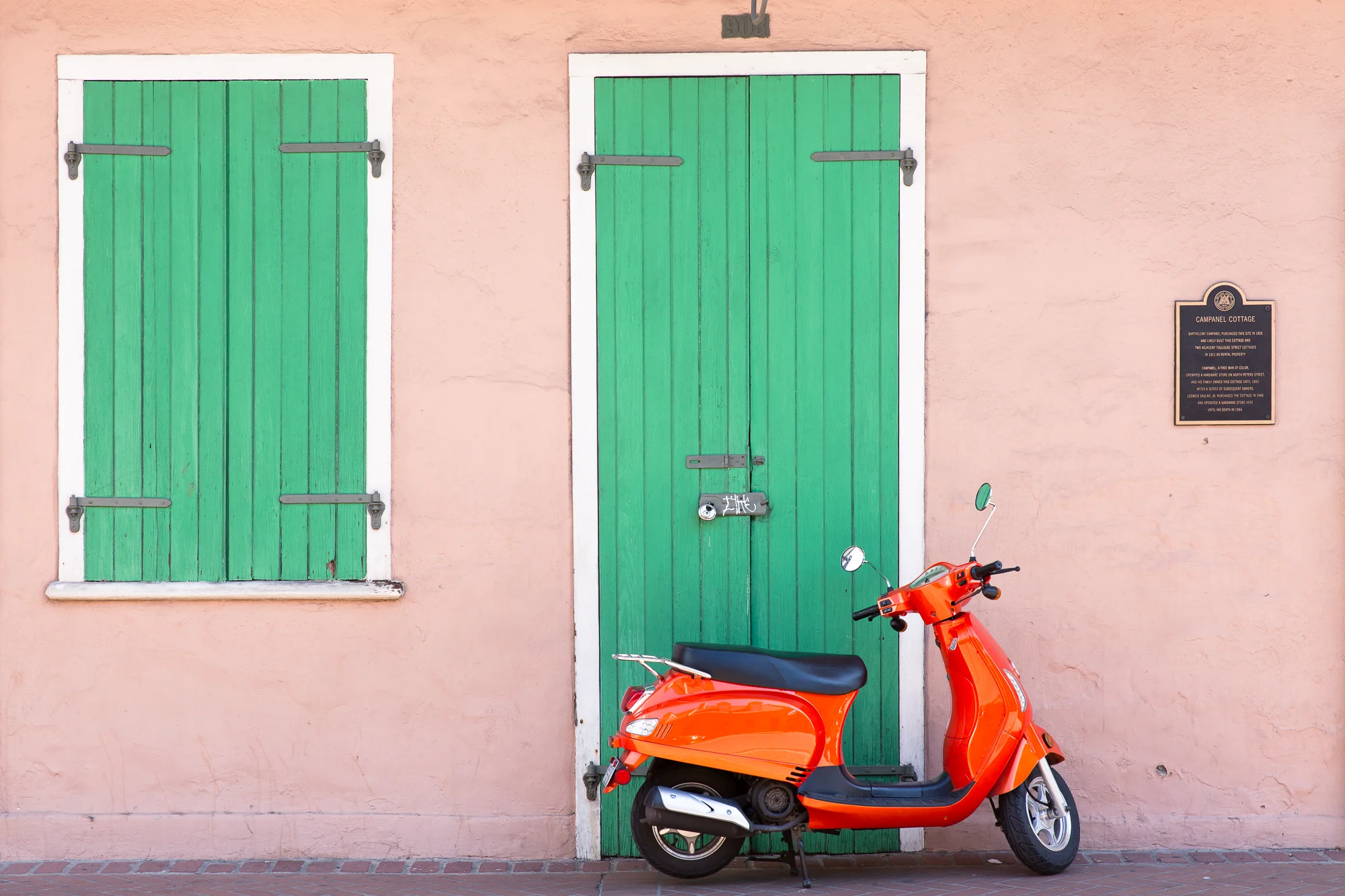 An orange scooter parked on brick paving against a pink stucco wall, beside a green plank door and a green louvered shutter trimmed in white. A small dark plaque hangs on the wall to the right. In color.