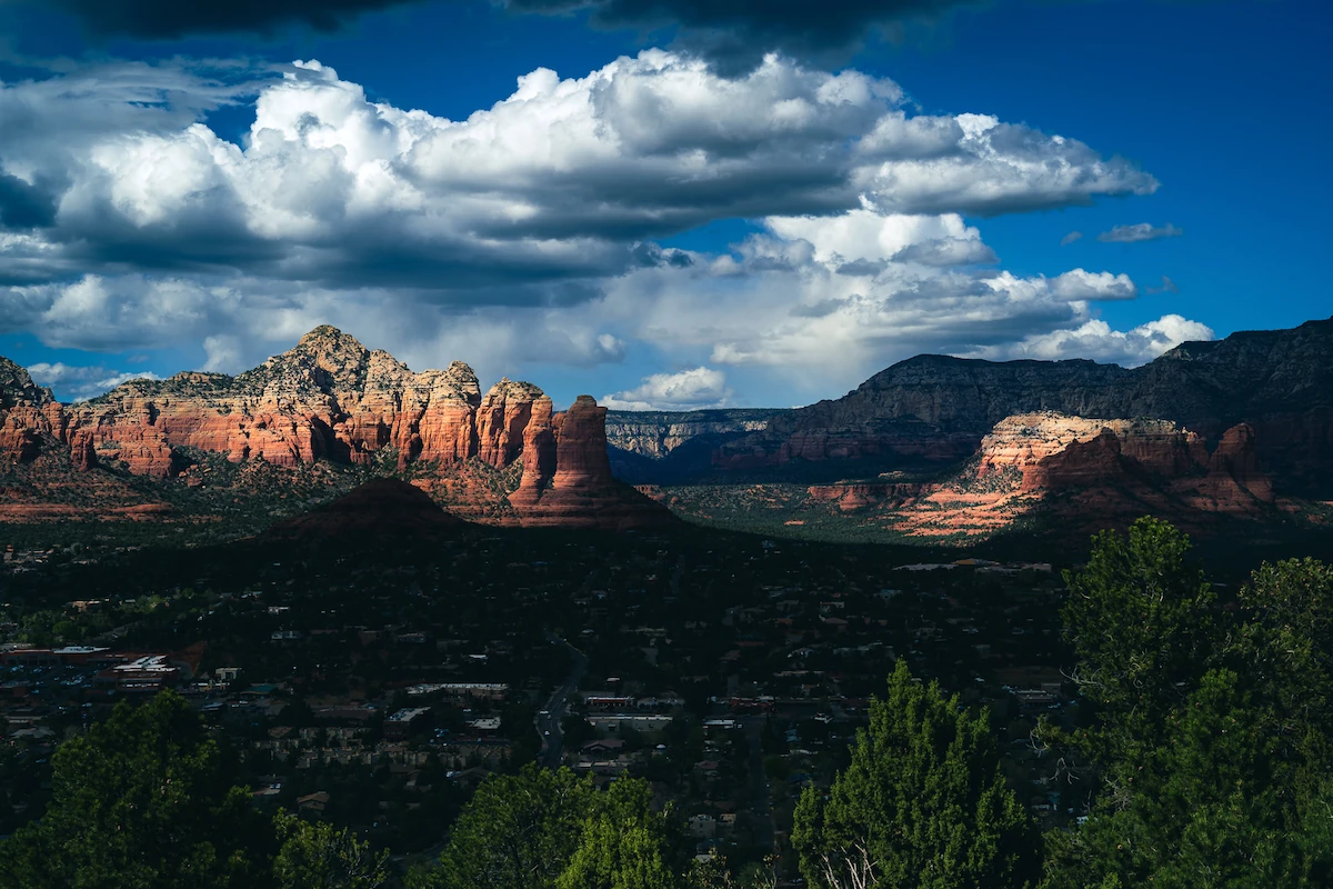 The red rock formations north of Sedona, Arizona, seen from above the town. Late afternoon sun catches the sandstone buttes and a tall spire at their center, while piled cumulus clouds throw the foreground ridges and the houses below into shadow.