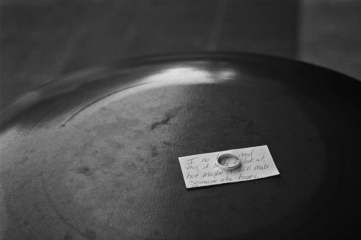 A wedding ring resting on a handwritten note, left on the lid of a trash can and lit from one side. The note reads: I no longer need this, it hurts to look at, but maybe it will make someone else happy. Black and white.