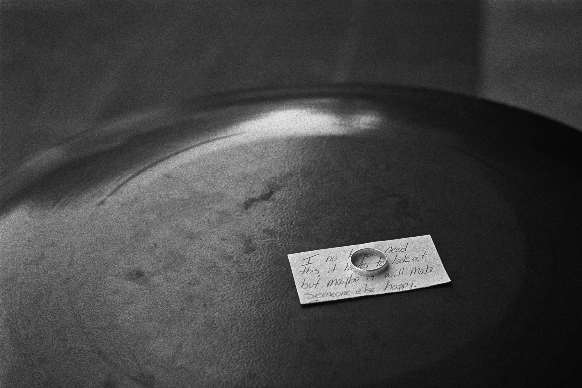 A wedding ring resting on a handwritten note, left on the lid of a trash can and lit from one side. The note reads: I no longer need this, it hurts to look at, but maybe it will make someone else happy. Black and white.