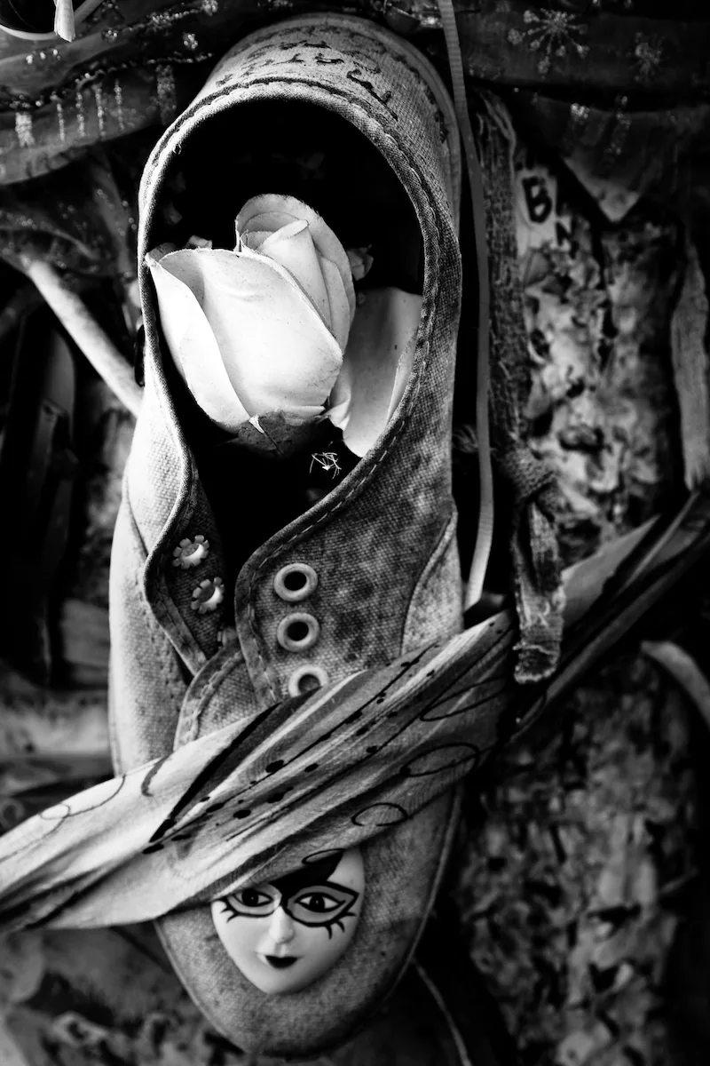 A white rose tucked into the mouth of a worn canvas sneaker fixed to a weathered wall, laces and eyelets close in the frame. Below it a doll's face looks out from under a painted brim. Black and white.