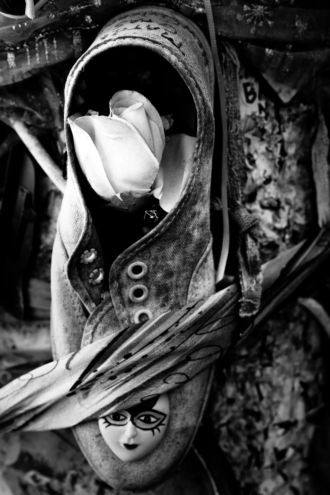 A white rose tucked into the mouth of a worn canvas sneaker fixed to a weathered wall, laces and eyelets close in the frame. Below it a doll's face looks out from under a painted brim. Black and white.